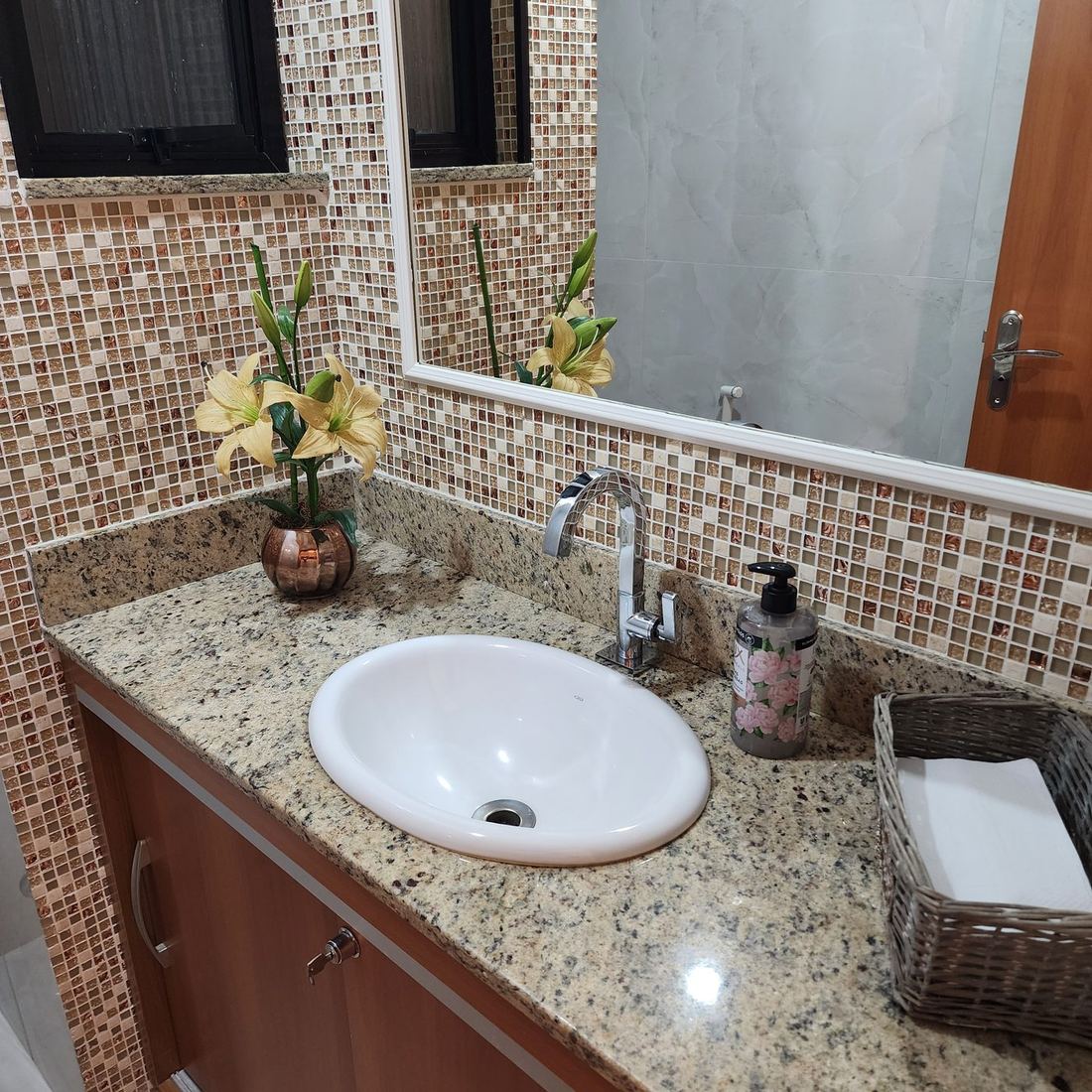 Banheiro com bancada de granito, pastilhas de vidro na parede e um arranjo de flores