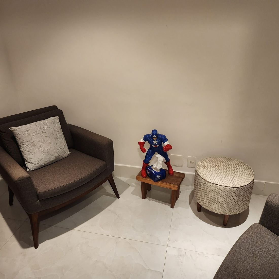 Sala de atendimento com duas poltronas cinza, pufe redondo e uma caixa de lenços sobre um banquinho de madeira